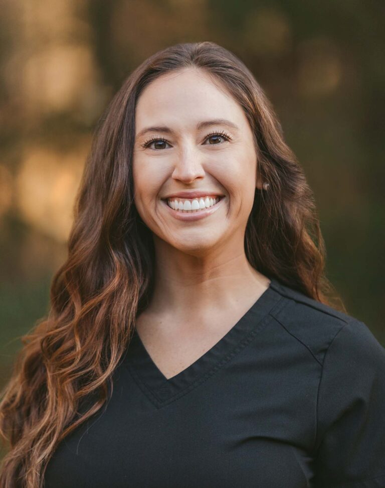 Headshot of Ashley wearing a black jacket with Dinwiddie Dental logo on the right chest, the background is outdoors.