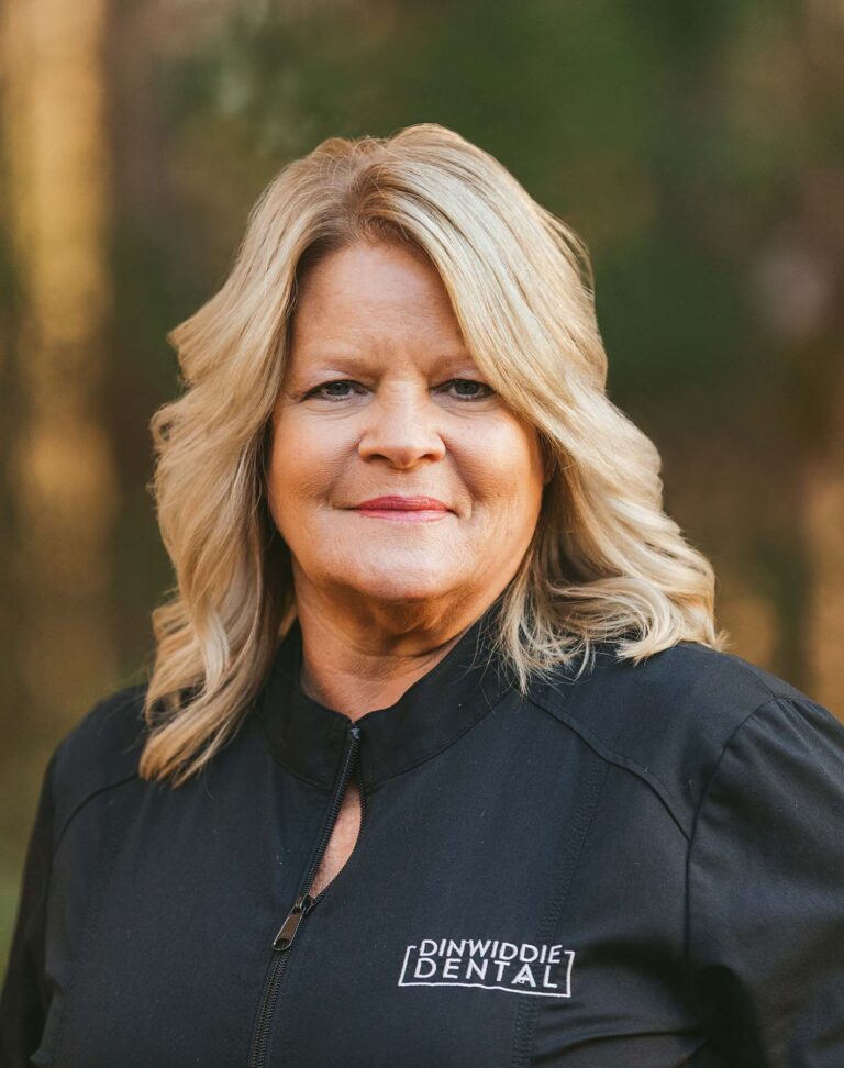 Headshot of Kim wearing a black jacket with Dinwiddie Dental logo on the right chest, the background is outdoors.