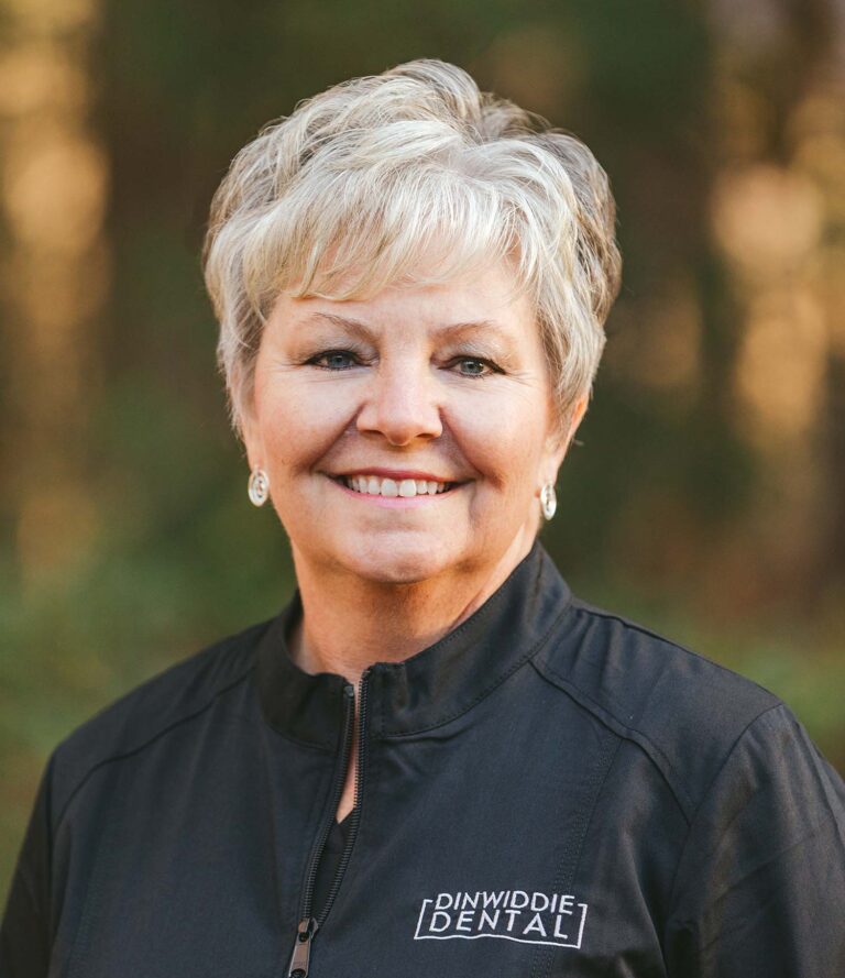 Headshot of Lori wearing a black jacket with Dinwiddie Dental logo on the right chest, the background is outdoors.