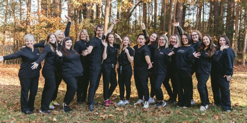 A group of people dressed in black poses playfully in a wooded area with autumn foliage. They raise their arms in various directions, creating a dynamic and lively scene.