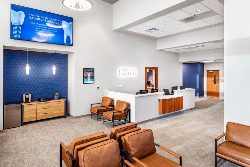 Modern dental office waiting area with brown leather chairs, a white reception desk, and a refreshment station beside a blue geometric wall. A screen promotes dental implants with benefits listed under the Dinwiddie Dental.