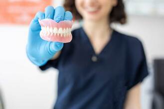 Person in dark blue medical uniform and disposable gloves holding set of artificial dentures with upper and lower teeth on pink bases, blurred background.